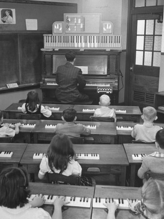 Black and white photograph of a classroom: seven children sit at keyboards; a male teacher in a suit plays a large keyboard and keys and notes on a staff light up above his head; to the left is a blackboard, and to the right is a door with nine panes of glass in the upper half; the middle pane is covered with a piece of paper drawn with a brick pattern