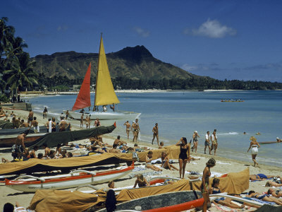 People Sunbathe and Walk on Sand Past Beached Outrigger Canoes