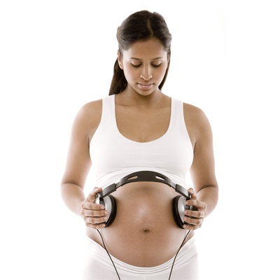 Pregnant woman dressed in white tank top and white pants holds noise-canceling headphones against her abdomen