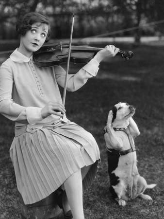 black and white photograph, woman with quizzical expression in 1920s day dress plays violin, brown and white dog sits up with its front paws over its ears