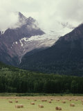 Buy Glacier Descending from Clouded Mountains Contrasts with a Field Dotted with Rolls of Hay at AllPosters.com