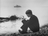 Actress Mia Farrow Pensively Sitting on Rocky Shore of Lake Geneva as Passenger Boat Passes By