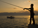 A Man Fishing from the Shore and a Fishing Boat in Water at Sunset