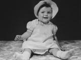 Baby Girl (12-18 Months) in Bonnet Sitting on Bedspread, Close-Up
