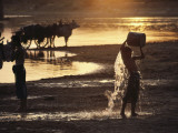 A Boy Pours a Bucket of Water over Himself at the Edge of a Lake