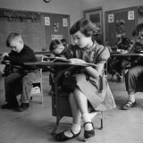 Cute Little Girl Busily at Work  Sitting in a Desk Chair in a Schoolroom  Other Pupils at Work Too