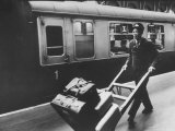 Railroad Porter Pushing Hand Truck Loaded with Baggage Past Passenger Car at Railroad Platform