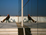 Man on Bench Reflected in Mirrored Window of Building Along Grado Promenade Near Town Centre