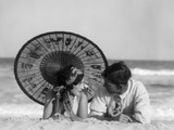 1920s Romantic Couple Looking At One Another Laying Face To Face Under Parasol On Sandy Beach