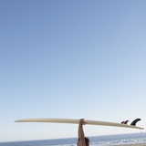 Head and arms of man carrying surf board above head at beach